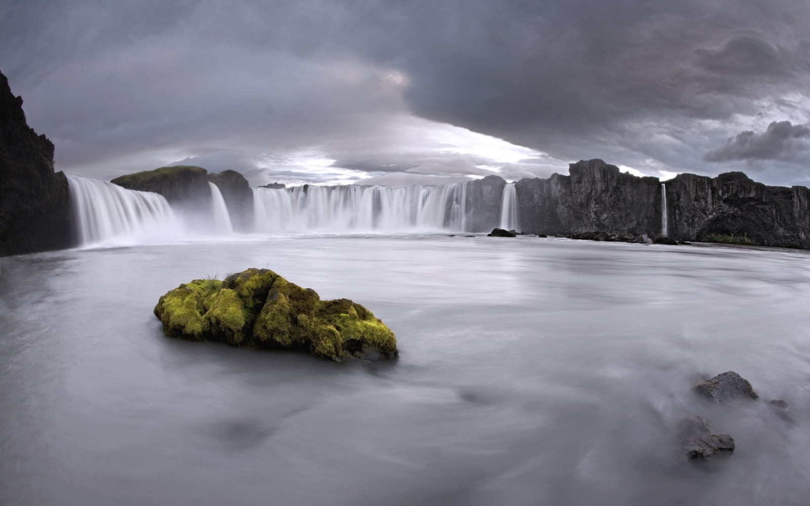 Uma cachoeira com uma pedra em primeiro plano e um céu escuro (seljalandsfoss, cachoeira, corpo de água, natureza, recursos hídricos)