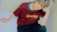 Choi Beomgyu en una camiseta casual color burdeos de Gola, sentado pensativo contra una pared.