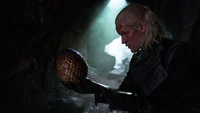 Daemon Targaryen segurando um ovo de dragão em uma caverna mal iluminada