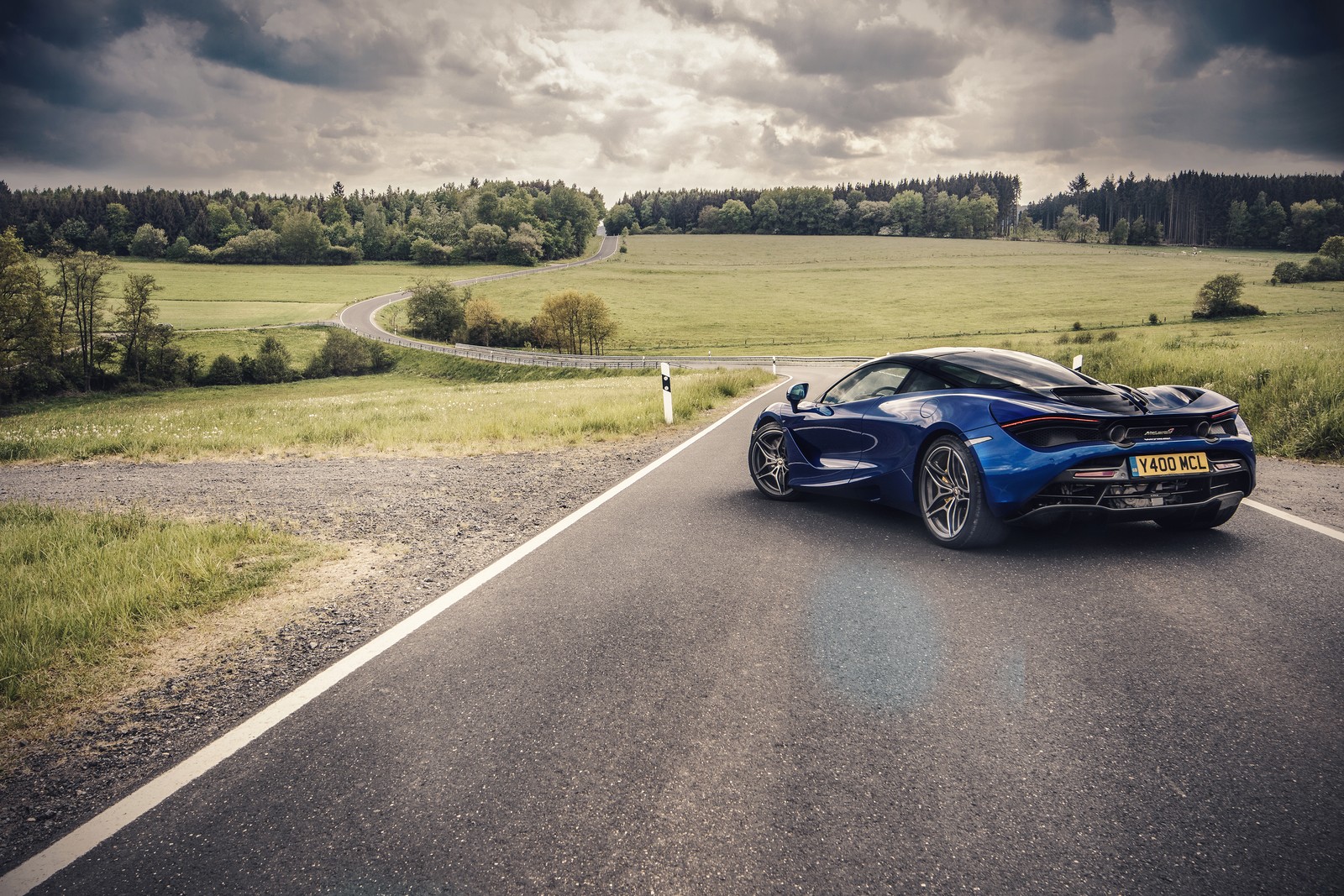 A blue sports car driving down a country road with a bird on the roof (road, mclaren 12c, mclaren automotive, sports car, car)