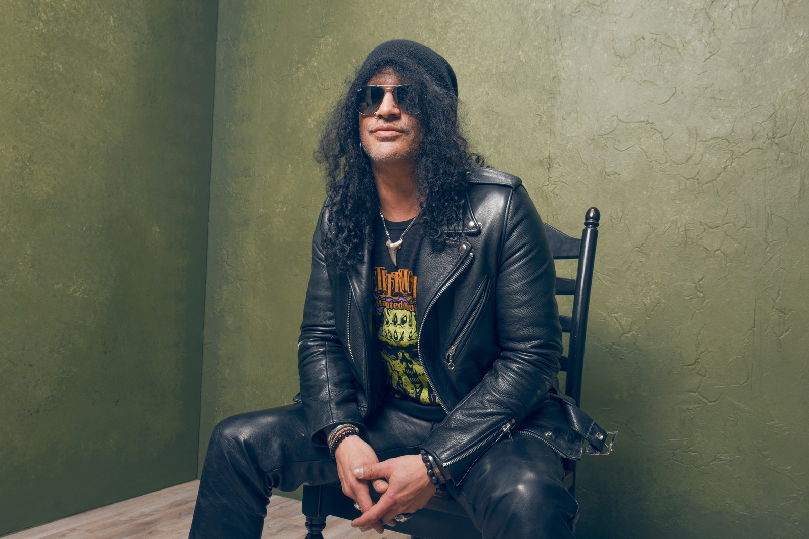 Um homem de jaqueta de couro e óculos escuros sentado em uma cadeira (slash, guns n roses, jaqueta de couro, jaqueta, legal)