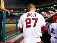 Baseball player in red jersey with 'Trout' and number 27, walking towards the dugout in a stadium.