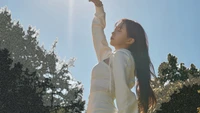 Haewon from NMIXX in a serene outdoor setting, reaching towards the sunlight, embodying grace and charm.