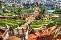 Vista aérea de un hermoso jardín botánico paisajístico con terrazas bien cuidadas, palmeras y vibrantes parterres de flores, con vistas a un paisaje urbano histórico y un templo destacado.