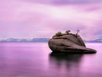 Exposition longue sereine d'un rocher solitaire entouré d'eaux tranquilles, avec un ciel rose époustouflant et des montagnes enneigées au loin au lac Tahoe.