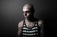 Monochrome portrait of a male model with intricate tattoos and skull-inspired makeup, set against a dark background.