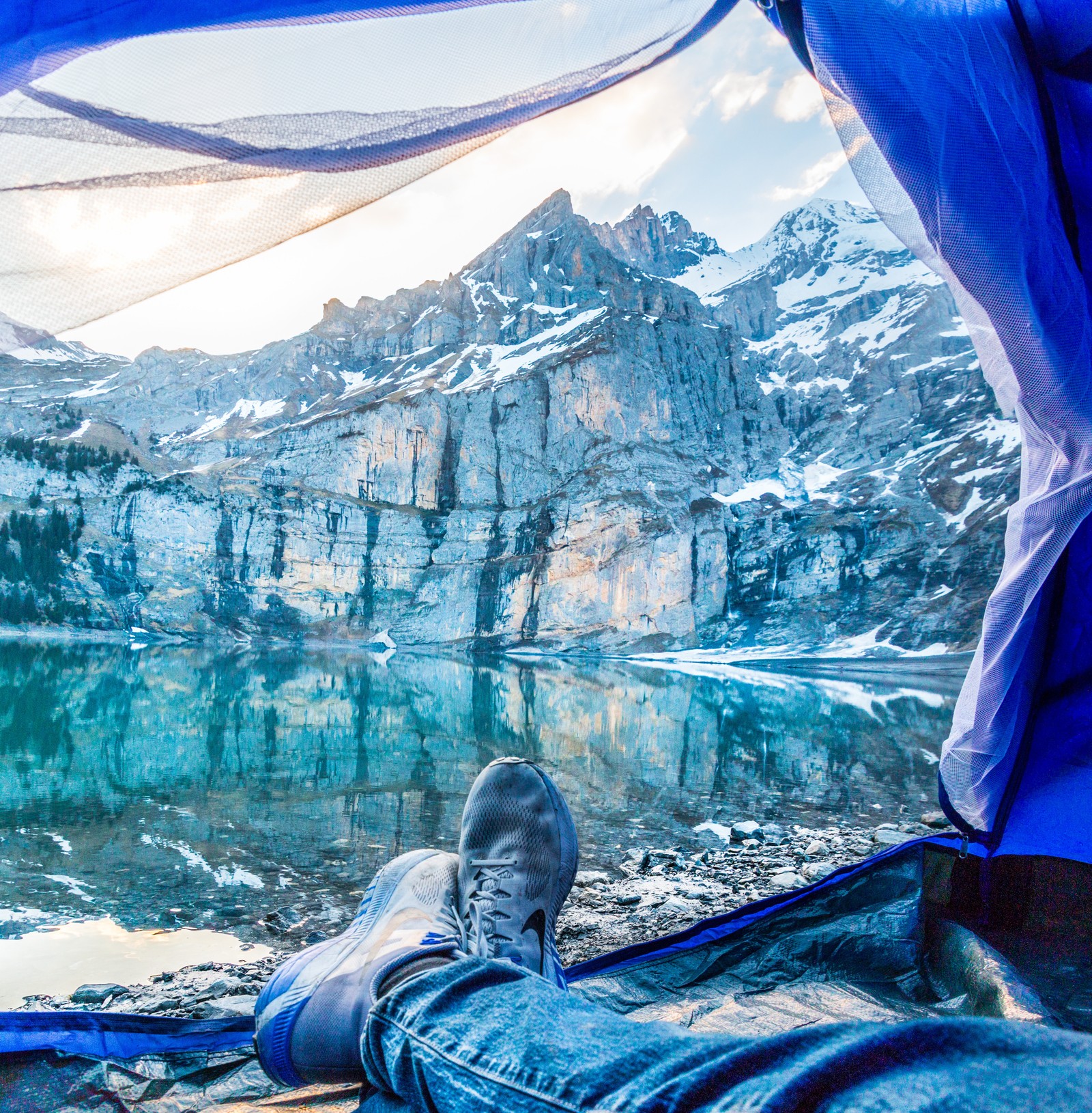 Alguien sentado en una tienda con los pies en alto y una montaña de fondo (tienda, campamento, azul, agua, forma glaciar)