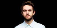 A young musician with styled hair and a well-groomed beard, wearing a white shirt against a dark background.