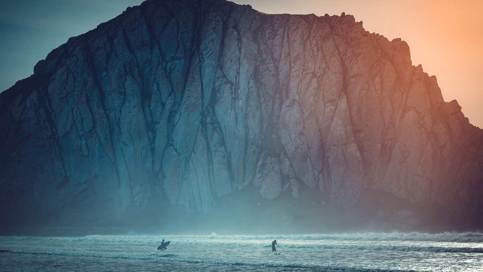 Los surfistas caminan por la playa frente a una gran roca. (agua, recursos hídricos, atmósfera, montaña, naturaleza)