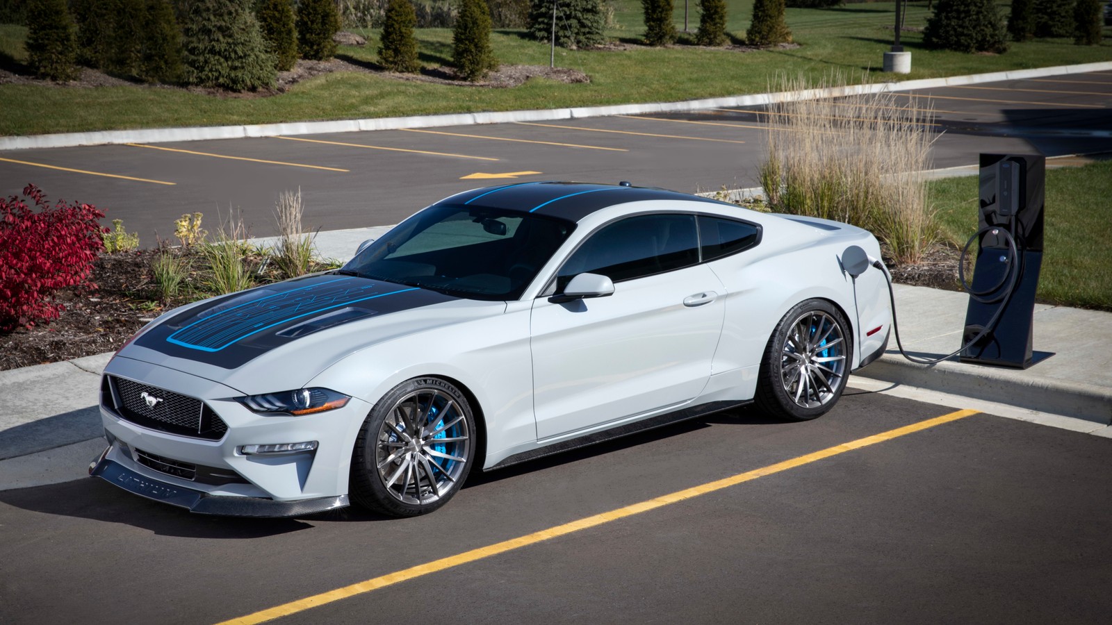 Une mustang blanche garée dans un parking à côté d'une station de recharge (voitures, ford motor company, ford, pony car, roue)