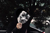 A person in a black hoodie holds a Guy Fawkes mask in front of their face, partially obscured by a dark tree backdrop, creating a striking contrast in a black and white scene.