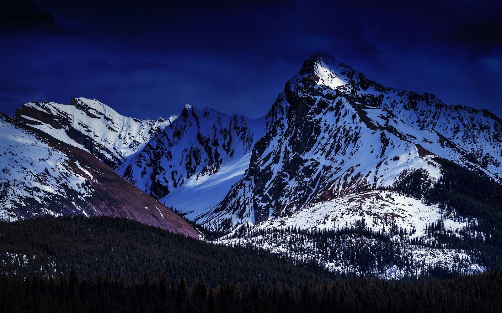 Vista de uma cadeia montanhosa com montanhas cobertas de neve ao fundo (montanhas glaciais, floresta, coberto de neve, céu escuro, árvores verdes)