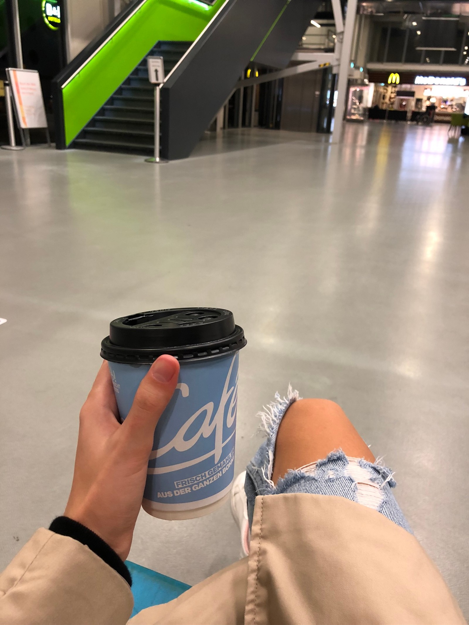 Alguien sostiene una taza de café en un centro comercial con una escalera verde (suelo, coches, ternero, aptitud física, pierna humana)