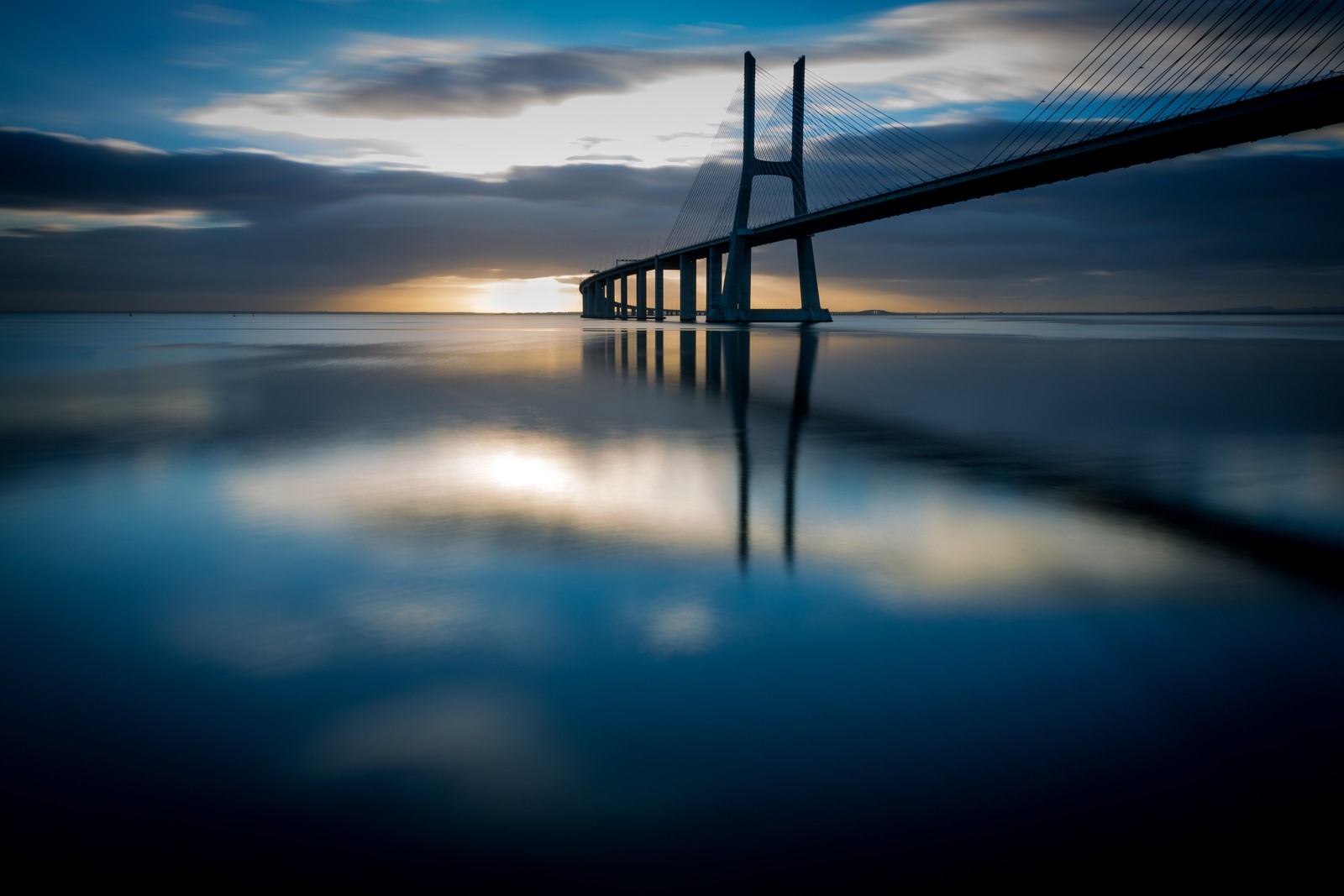 Une vue d'un pont sur un plan d'eau au coucher du soleil (lisbonne, lisbon, bleu, eau, réflexion)