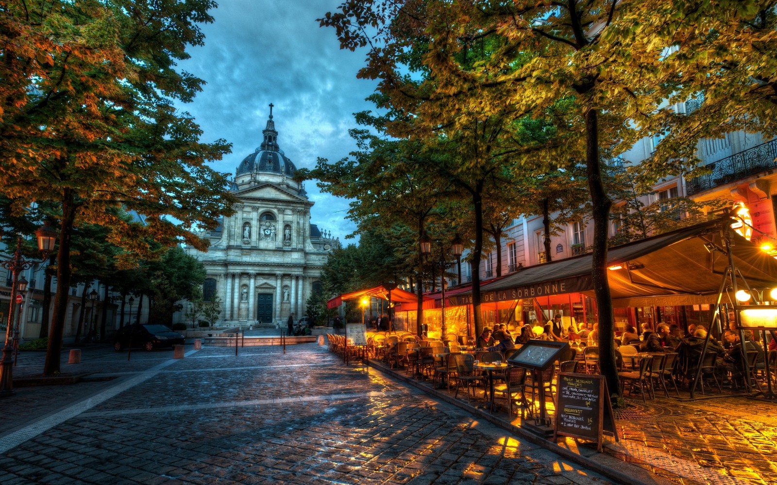 Arafed view of a street with tables and chairs and a church in the background (tree, landmark, town, autumn, night)