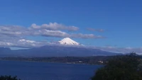 Snow-capped stratovolcano rises majestically over a tranquil lake, framed by a clear blue sky and clouds, surrounded by lush highland scenery.