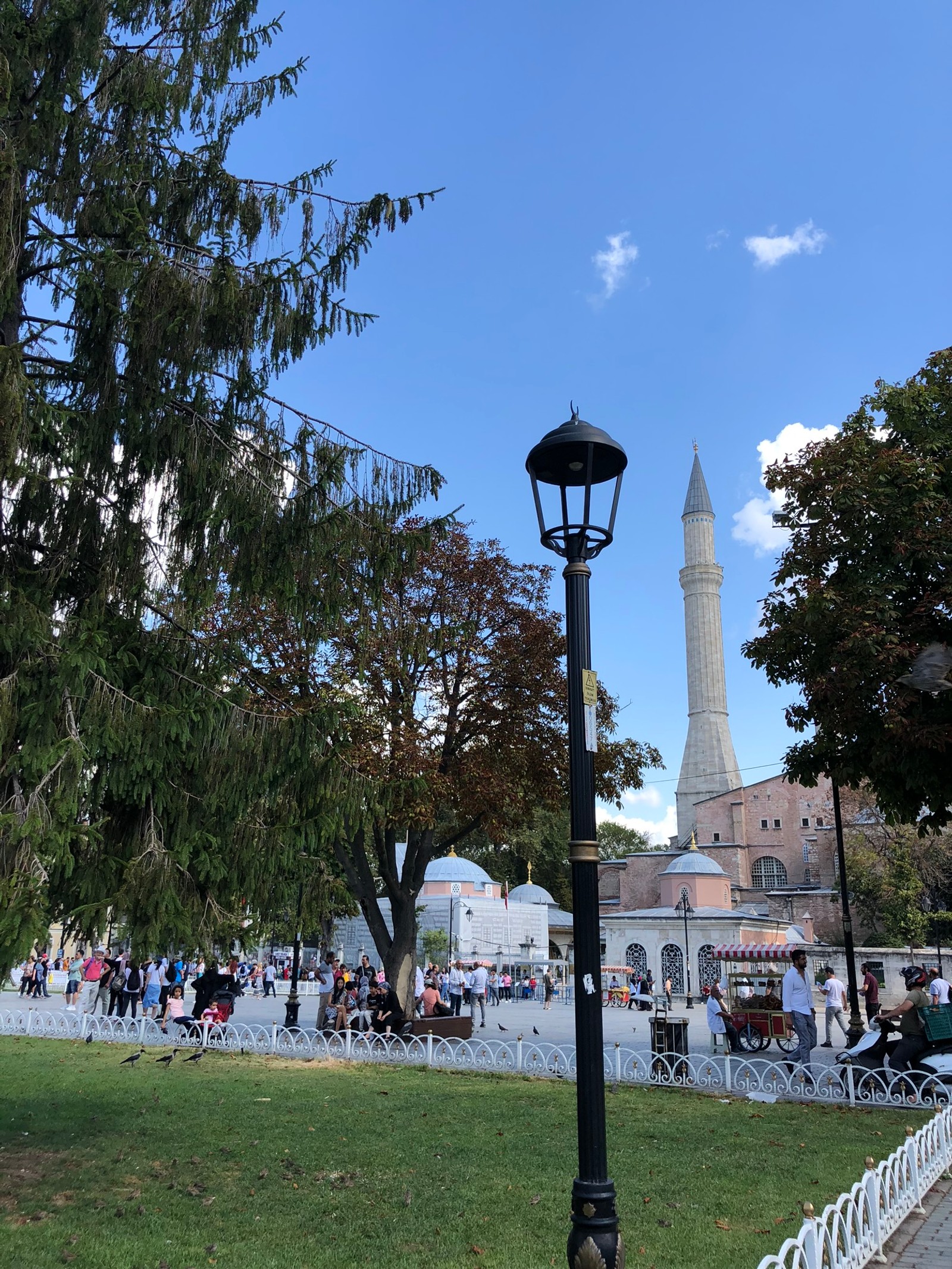 Il y a un lampadaire au milieu d'un parc avec des gens qui se promènent autour (tourisme, attraction touristique, réverbère, arbre, espace public)