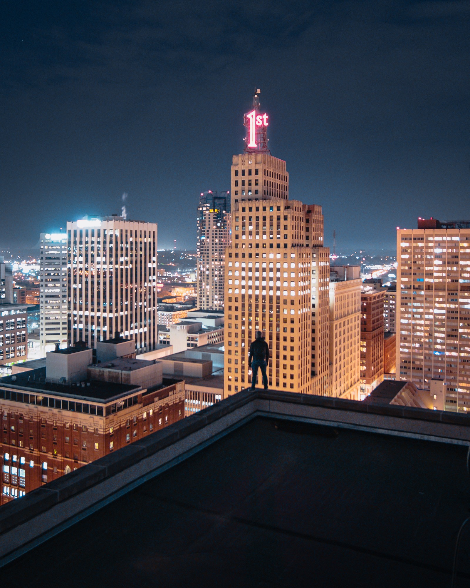 Vue nocturne d'un homme debout sur un toit surplombant une ville (bâtiment, gratte ciel, façade, établissement humain, ville)