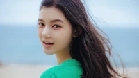 Danielle Marsh of NewJeans smiles at the camera against a beach backdrop, her hair gently flowing in the breeze.