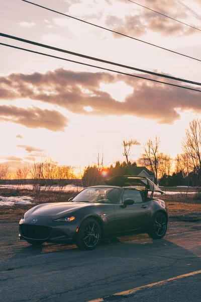 Voiture de sport Mazda MX-5 garée sur une route pittoresque au lever du soleil, encadrée par des arbres et un ciel nuageux.