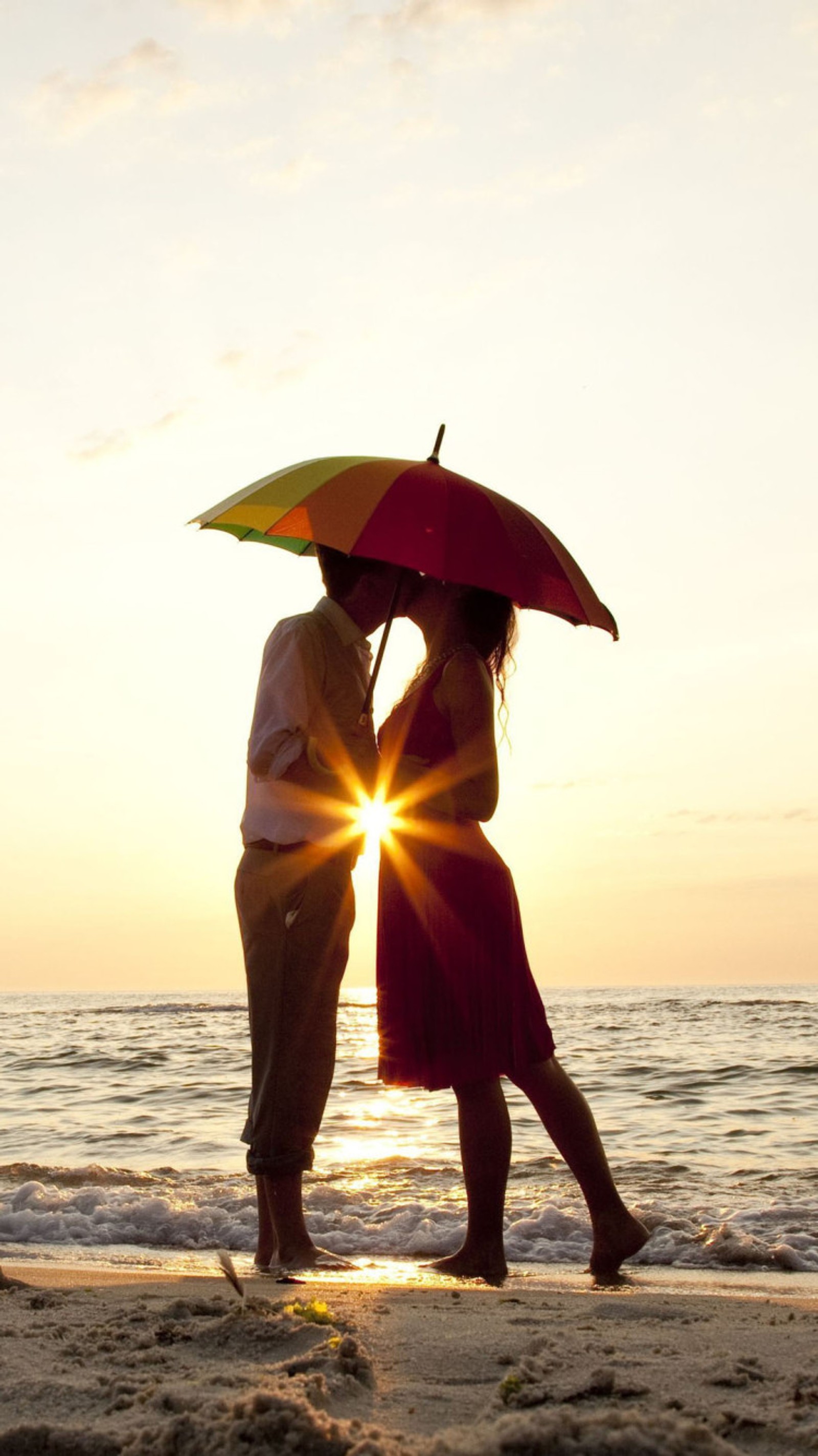 Ils se tiennent sur la plage sous un parapluie au coucher du soleil (couple, embrasser)