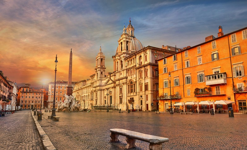 Arafed view of a city square with a bench and buildings (rome, city, town, town square, plaza)