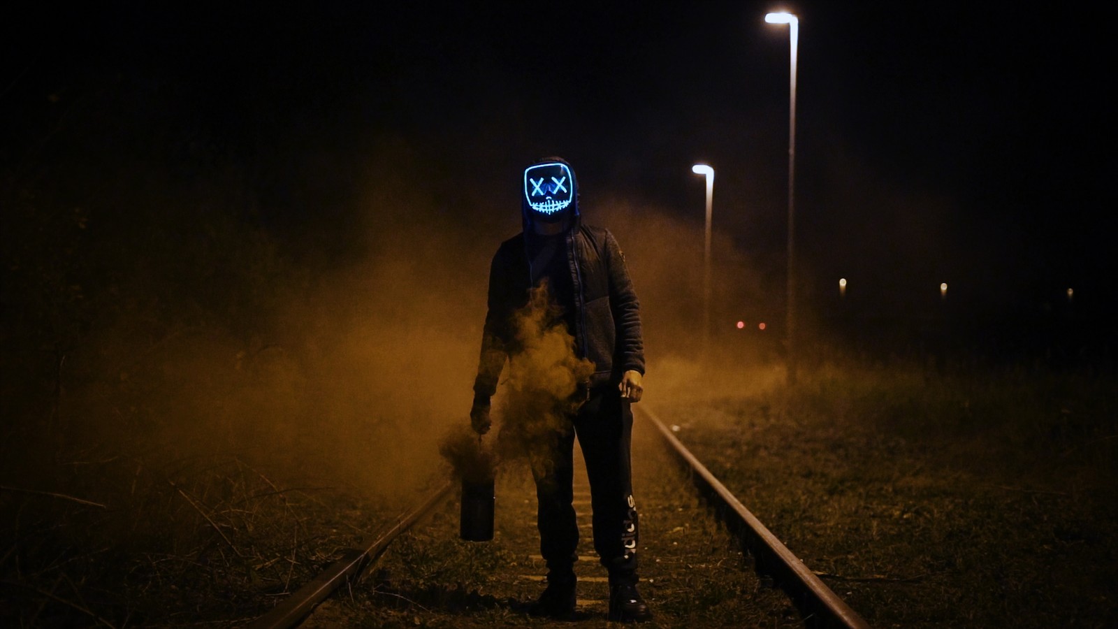 Um homem encapuzado com uma máscara neon em pé em uma linha de trem (máscara, escuridão, luz, iluminação, noite)