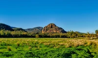 Campo vibrante com uma colina rochosa proeminente sob um céu azul claro, emoldurado por vegetação exuberante em um ambiente selvagem tranquilo.