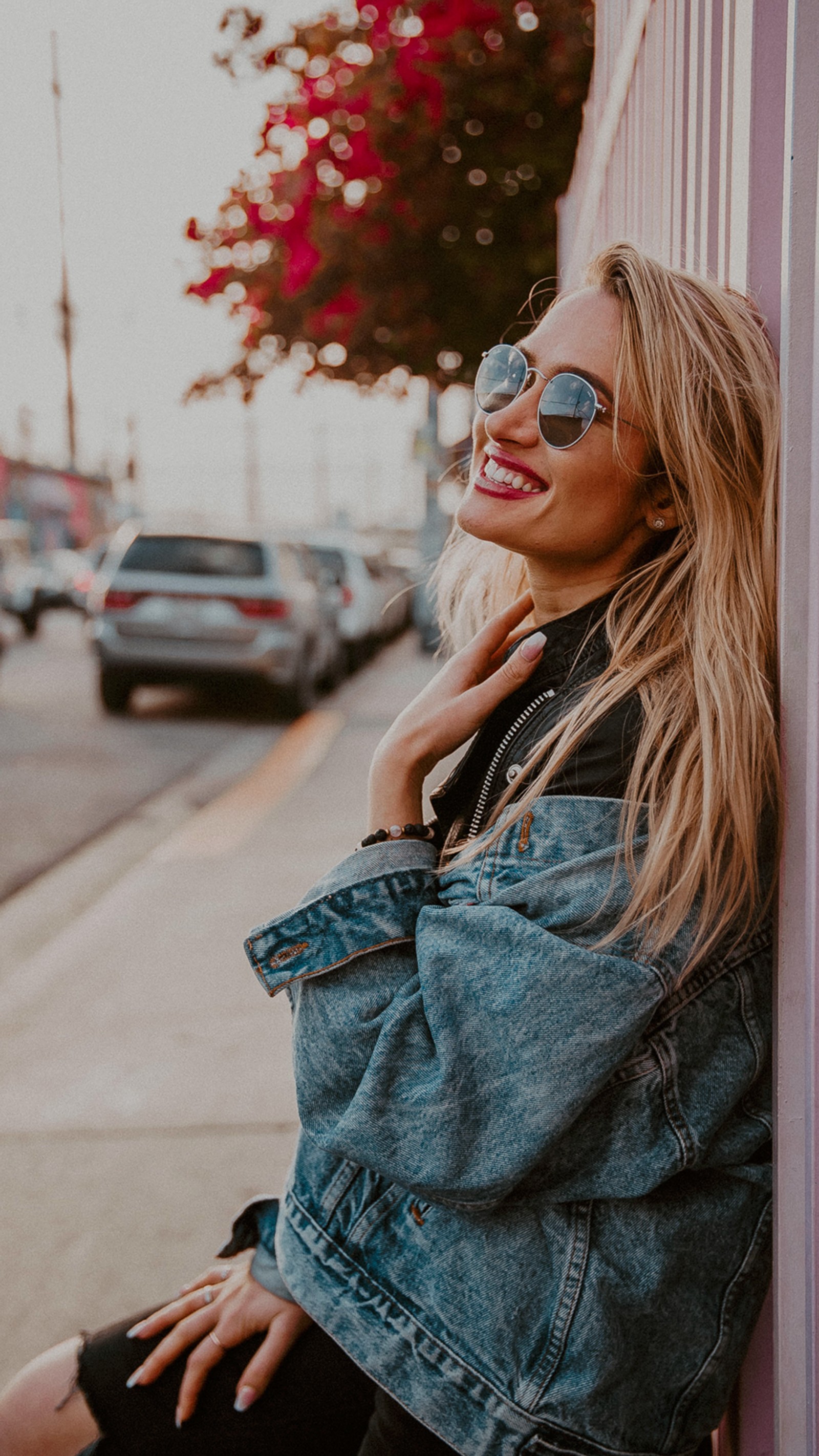 Eine blonde frau mit sonnenbrille, die an einer wand auf einer stadtstraße lehnt (sonnenbrillen, ios, lippe, oberbekleidung, schulter)