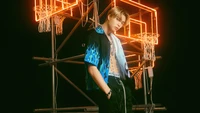 Johnny von NCT U strahlt Charisma in einem lebhaften blauen und schwarzen Outfit vor einem neonfarbenen Basketball-Hintergrund aus.