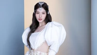 Minatozaki Sana em um elegante vestido branco com mangas bufantes dramáticas, adornado com uma tiara de joias, contra um fundo pastel.