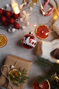 Eine festliche Anordnung mit einem Weihnachtsornament, getrockneten Orangenscheiben, einem verpackten Geschenk und dekorativen Akzenten, alles vor einem gemütlichen, strukturierten Hintergrund.