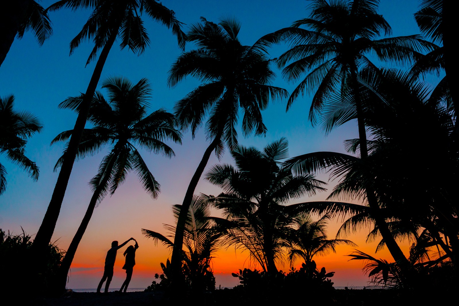 Silhouette von zwei personen, die am strand mit palmen stehen (paar, tropischer strand, romantisch, sonnenuntergang, dämmerung)