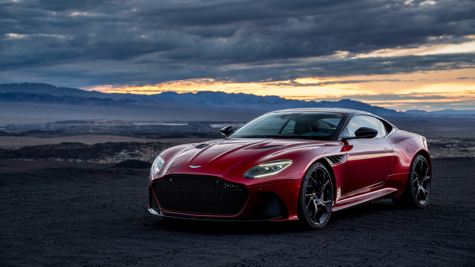 Un coche aston rojo estacionado en un camino de tierra frente a una montaña (aston martin, aston martin db11, aston martin vanquish, coche, coche deportivo)