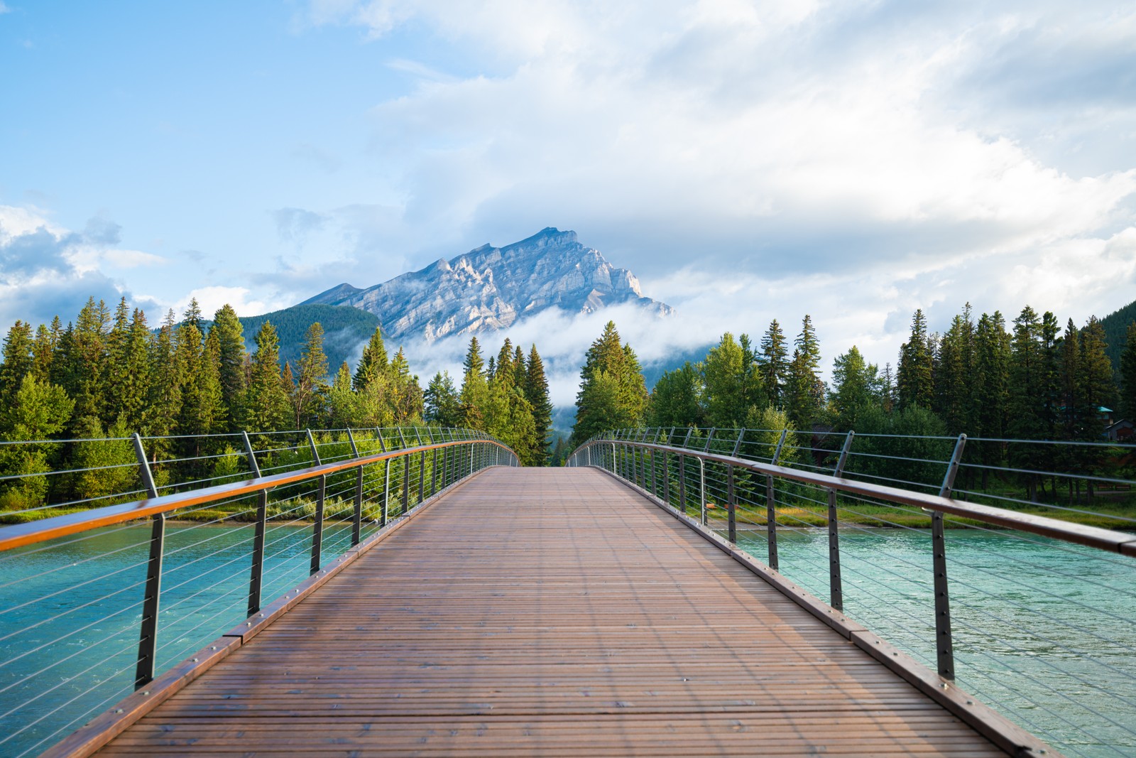 Uma ponte árabe sobre um rio com uma montanha ao fundo (ponte de madeira, parque nacional de banff, banff national park, árvores verdes, pico da montanha)