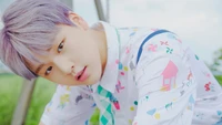 Nam Do Hyun dans une chemise colorée, mettant en valeur ses cheveux violets distinctifs contre un arrière-plan extérieur vibrant.