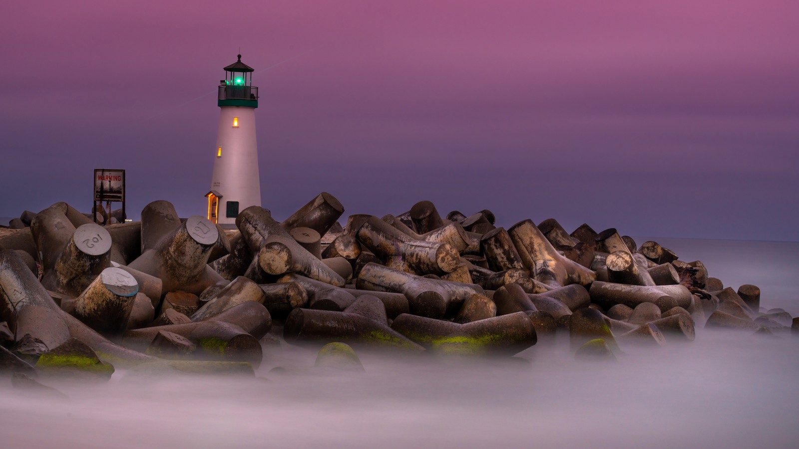 Farol arafed em uma praia rochosa com um céu roxo. (farol, água, torre, madeira, planta)