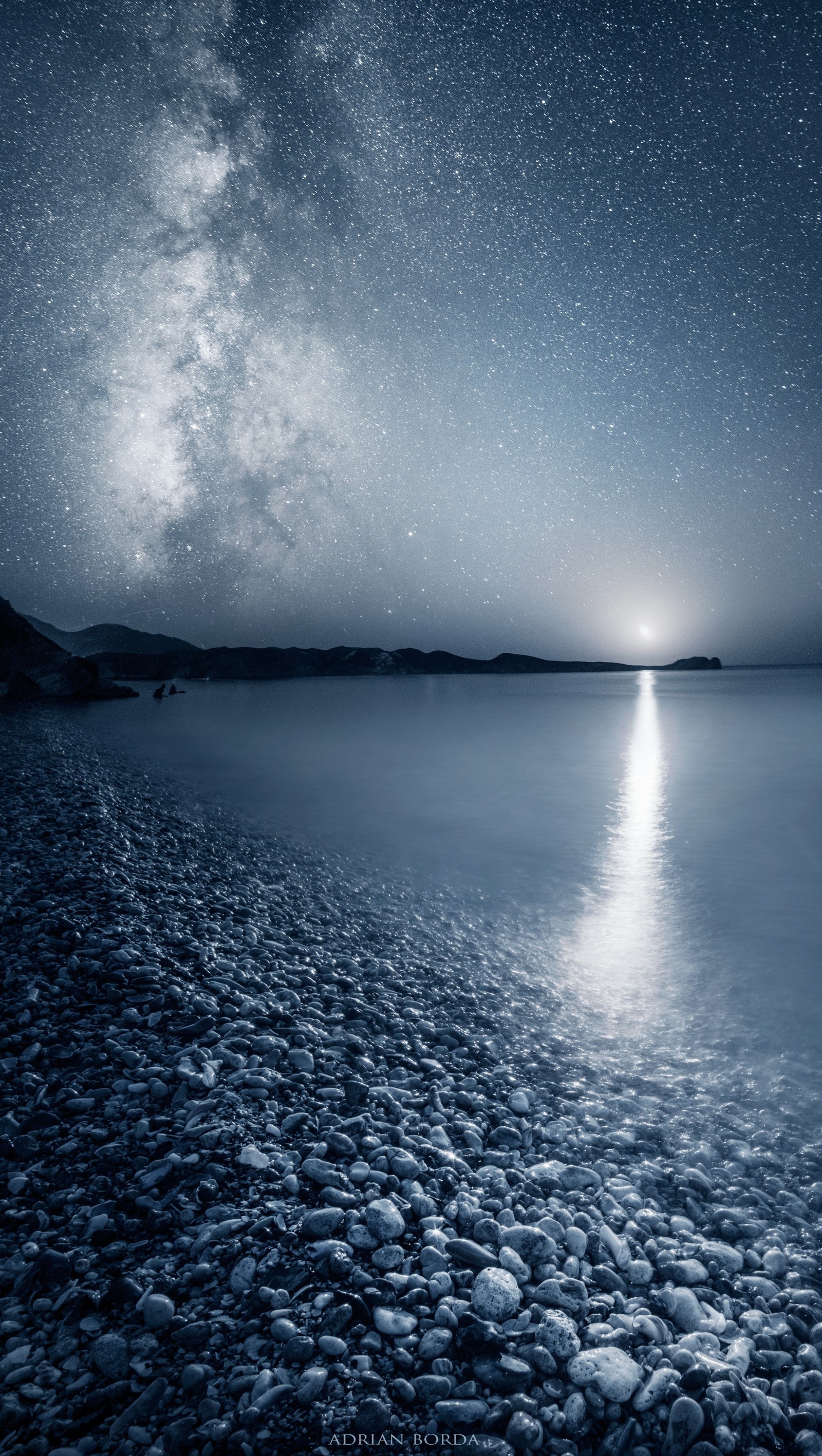 Céu estrelado à noite sobre uma praia com pedras e água (adriano, praia, borda, lua, noite)