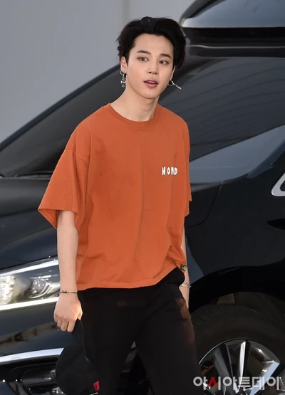 Jimin do BTS em uma camisa oversized laranja, caminhando confiantemente perto de um carro preto.