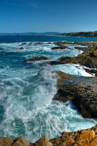 Des vagues dynamiques s'écrasent contre un promontoire rocheux sous un ciel bleu clair