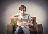 Jovem sentado em um sofá, comendo batatas fritas de uma tigela, com uma garrafa de cerveja próxima.