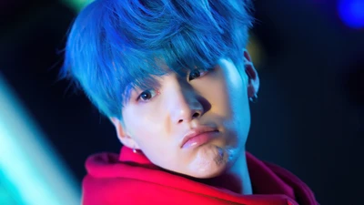 Suga from BTS with blue hair, wearing a red hoodie, captured in a dramatic lighting setup.