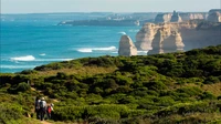 Wanderer durchqueren üppiges Terrain entlang der dramatischen Klippen der Zwölf Apostel, mit Blick auf das lebendige Meer im malerischen Naturreservat von Melbourne.